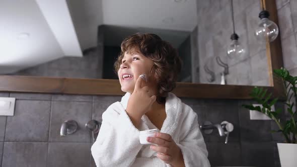 Little Boy in white Robe Sitting Near Bathroom Mirror and Playing with Skin Care Cream
