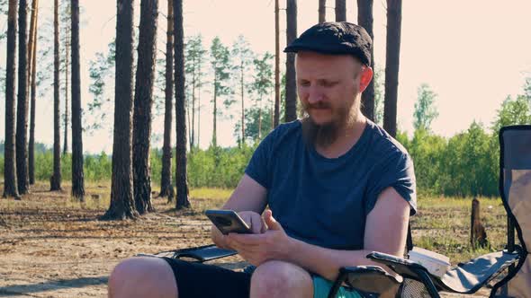 A Man with a Wellgroomed Appearance and a Graceful Beard in a Beautiful Summer Green Forest Sits in