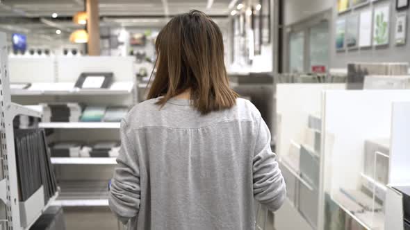 Young Asian woman rides shopping cart choosing new furniture in warehouse.