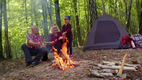 Senior Grandmother Grandfather with Granddaughter Cooking Frying Sausages Over Campfire in Wood