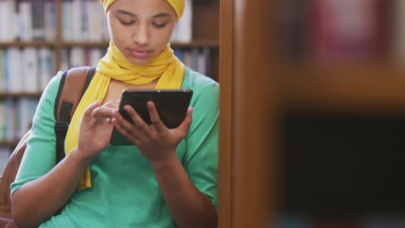 An Asian female student wearing a yellow hijab leaning against bookshelves and using tablet