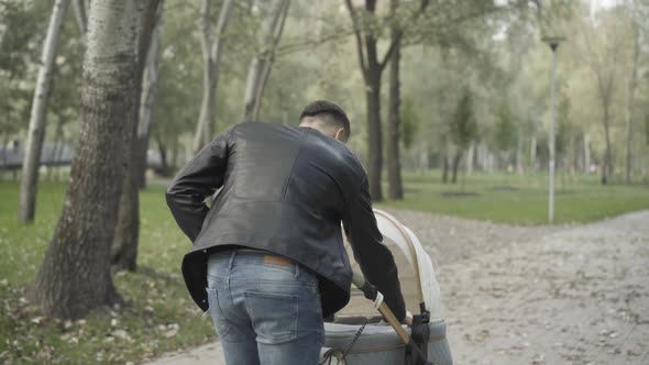 Back View of Young Brunette Caucasian Man Pushing Baby Stroller in Summer Park and Adjusting Blanket