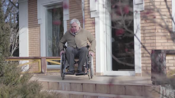 Old Caucasian Man in Wheelchair Rolling To Stairs Without Ramp Outdoors and Throwing Up Hands in