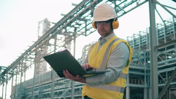 Engineer in Safety Wear is Observing Oil Refinery and Using a Laptop