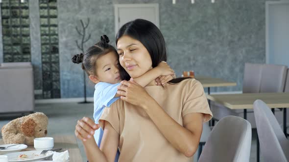 Little Child Girl Hugs Mum Brunette with Smile Near Table