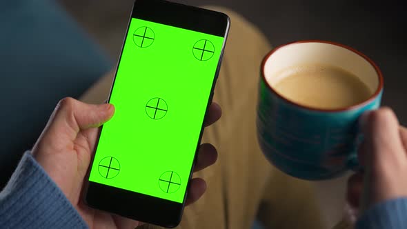 Man Using Smartphone with Green Mockup Screen in Vertical Mode and Drinking Coffee