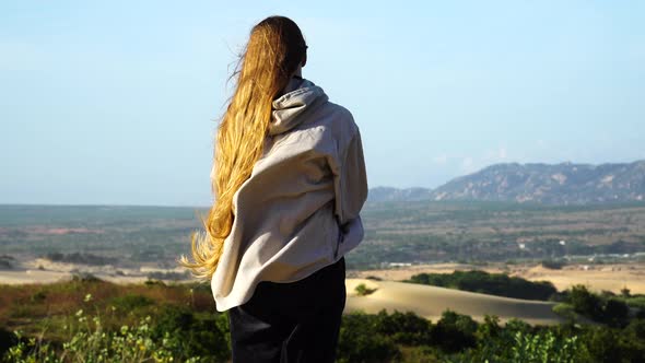 Woman from behind with blond long hair and shirt blowing In the wind standing at viewpoint of scenic