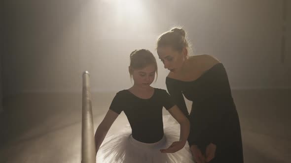 Medium Shot of Slim Woman Explaining Third Ballet Position Talking with Girl in Dance Studio Backlit