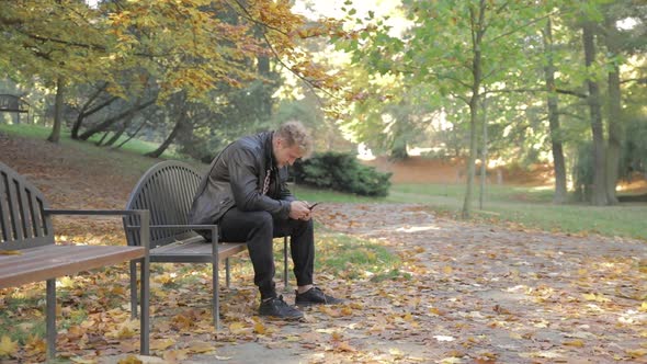 A young man looking at his mobile phone on a park bench alone on at autumn day