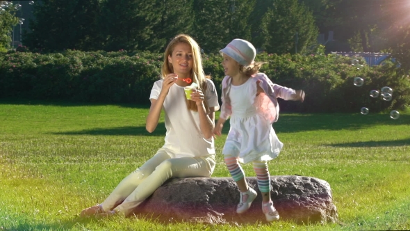 Young Mother and Her Daughter Playing with Soap Bubbles in the City Park