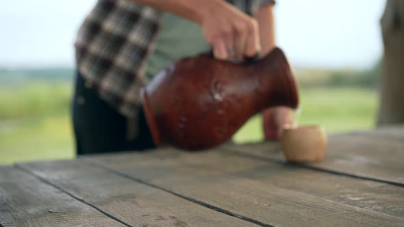 Blurred Unrecognizable Man Pouring Milk From Clay Pot in Cup with Female Hand Putting Dairy Bottle