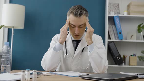 Tired senior male doctor sitting alone in the office in front of a laptop and closed it