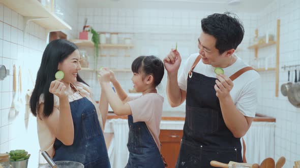 Asian happy family parent with young kid girl holding cucumber pieces in front of eye look at camera