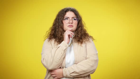 Pensive Young Plussize Woman in Eyeglasses Standing at Yellow Background Thinking