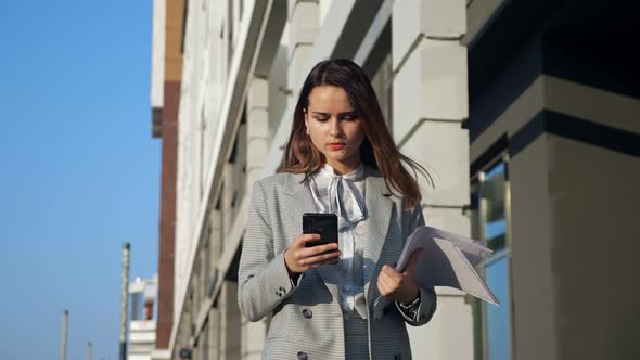 Young Woman in a Business Suit with a Phone and Documents Walking Down the Street