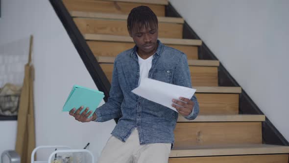 Young Sad African American Man Comparing Online Information on Tablet and Graph Standing in Home