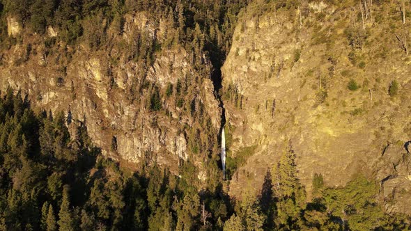 Aerial lowering over Corbata Blanca waterfall hiding between steep mountains with pine trees at suns