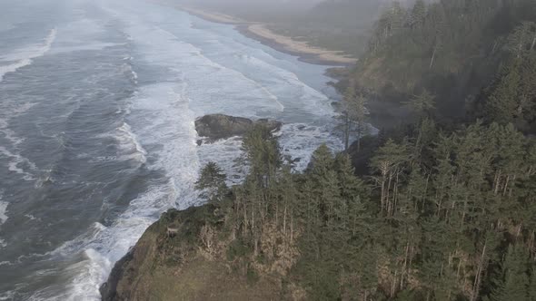 Passing over a rugged tree covered coastal outcrop, light mist, crashing waves, aerial