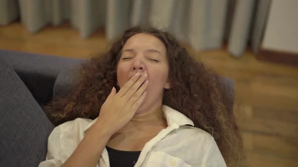 Sleepy Mixed Race Young Woman Yawns Lying on Grey Sofa