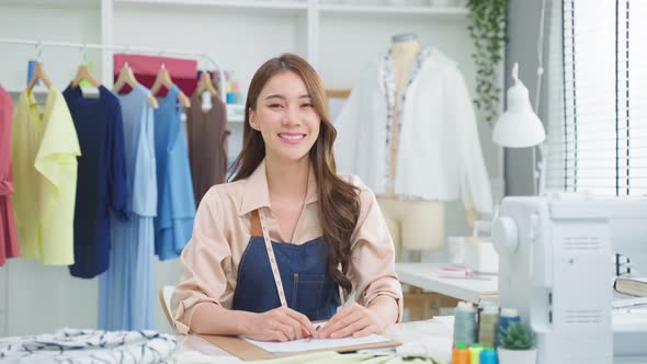 Portrait of Asian young tailor woman working on clothes in tailoring atelier and looking at camera.