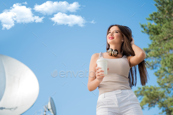  around her neck, holding takeaway coffee and posing against background of parabolic satellite dishes that receive wireless signals from satellites.
