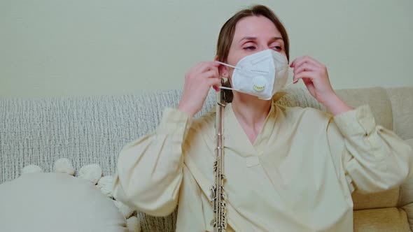 Woman musician with flute in medical face mask playing at home on sofa in living room