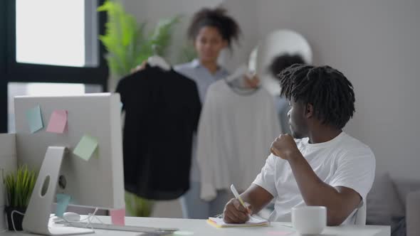 Thoughtful Man Writing Business Idea with Pen in Sketchpad As Woman Choosing Clothes at Background