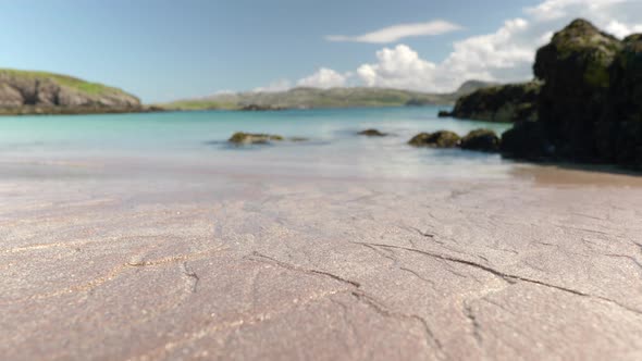 Close up shot focused on a sandy beach in the foreground as water flows across a beach into a turquo