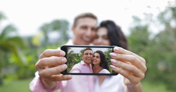 Couple Hold Cell Smart Phone Take Selfie Photo Embracing Kissing Outdoors Over Tropical Forest