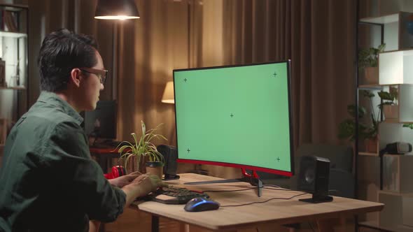 Asian Male Works With Mock Up Green Screen Computer Display. He Works At Night