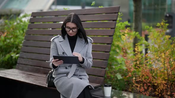 a Brunette in a Gray Coat Glasses and a Paper Cup is Sitting on a Wooden Bench with a High Back and