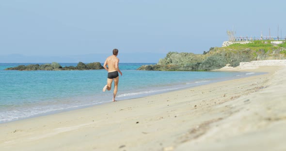 The Handsome Man with a Perfect Athletic Body in Swimming Trunks Having Fun on a Deserted Beach in