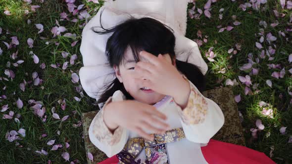 Korean Girl Child in a National Costume Lies on the Back in a Garden with Cherry Blossoms in Spring
