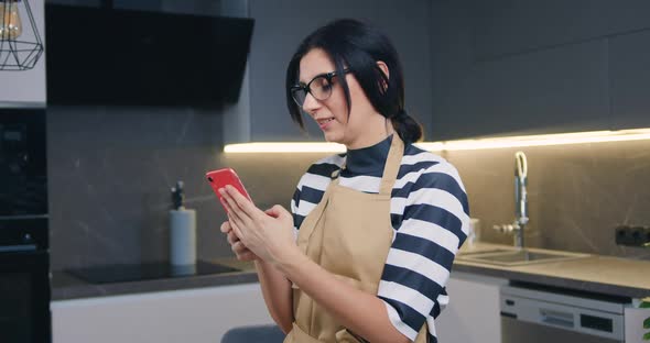 Woman in Glasses Standing in the Cozy Kitchen and Typing Sms on Mobile to Her Friend