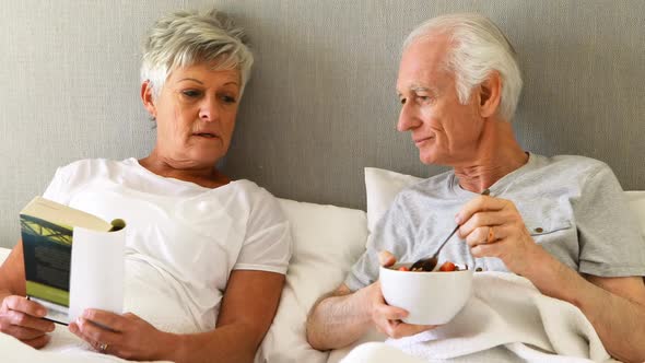 Senior man having breakfast while woman reading a book on bed