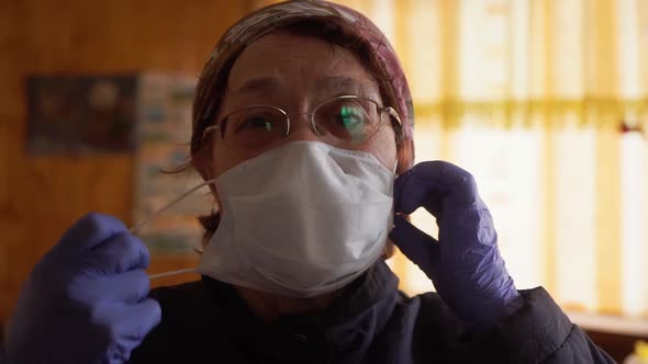 An Elderly Woman Puts Off a Protective Medical Mask Near the Window, Looks at the Camera. COVID-19