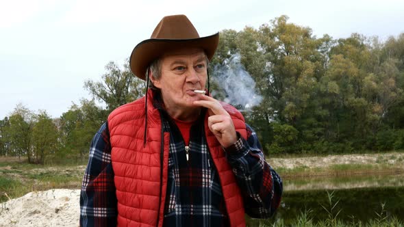 Mature Adult Man in Cowboy Hat and Clothes, Set Fire Cigarette From Lighter and Inhales Smoke.