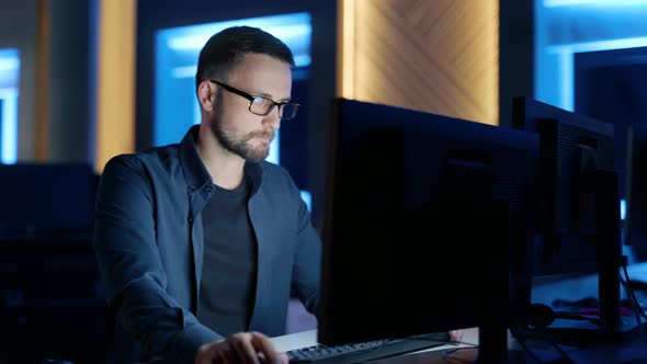 A Man with Glasses Sits and Thinks Typing on a Computer in the Office at Night