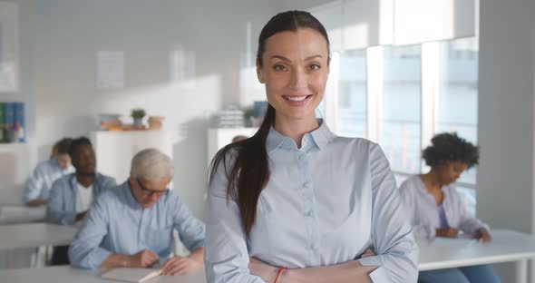 Portrait of Young Woman Teacher Smiling at Camera with Adult Students Sitting at Desk on Background