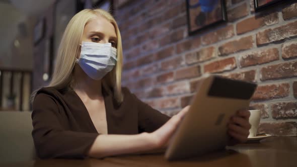 Attractive Young Woman in a Medical Mask Scrolls the Tablet in a Cafe