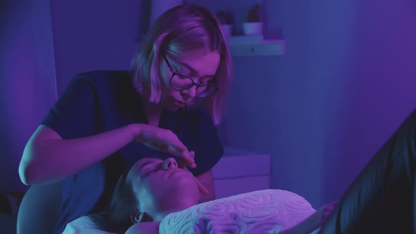 Blonde Young Woman Massagist Doing a Face Massage Using Her Fingers on Her Female Client Dark Purple