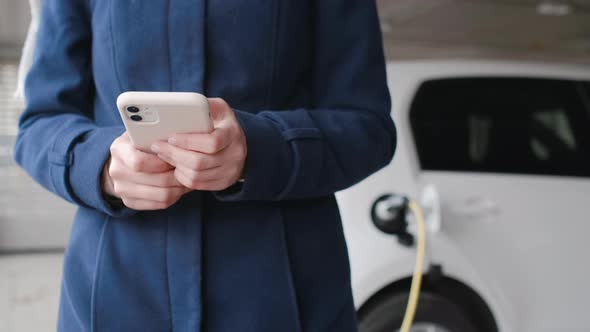 Woman Using Her Mobile Phone on the Background of the Charging Electric Car at the Parking