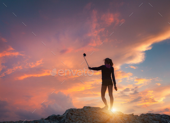 t sunset. Landscape with girl is photographing herself on the mountain peak against colorful sky with orange and red clouds. Lifestyle background