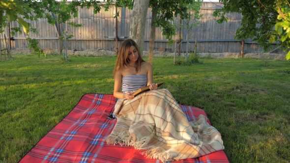 Portrait of a Cute Girl Reading a Book, Sitting on a Blanket Under a Tree