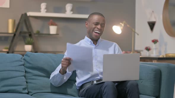 African Man with Laptop Reacting to Loss on Documents Sofa