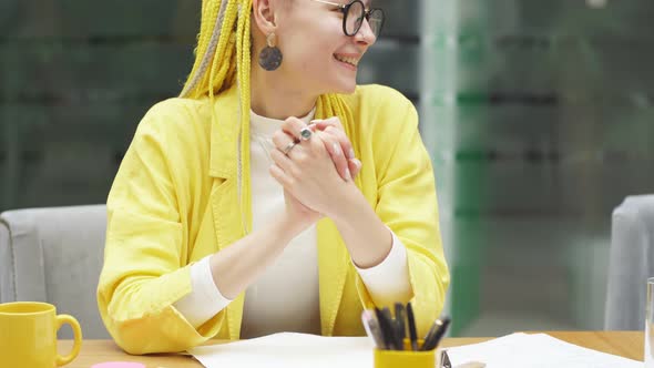 Original Cute Girl in Glasses with Yellow Pigtails Smiles Sitting at the Desk in the Office