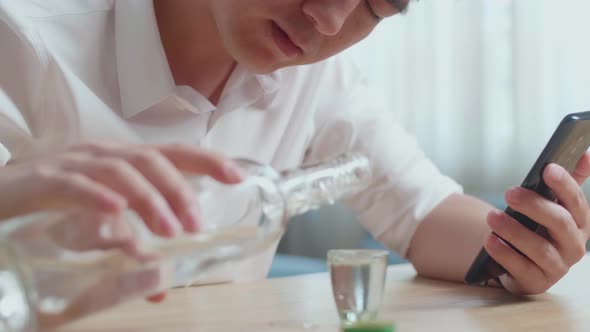 Close Up Of Drunk, Depressed Asian Man Pouring Vodka Before Drinking While Using Smartphone