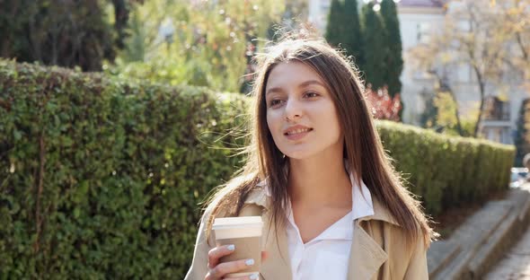 Young Attractive Business Woman Walks Around Historic Green Urban Area, with Take Away Cup and Drink