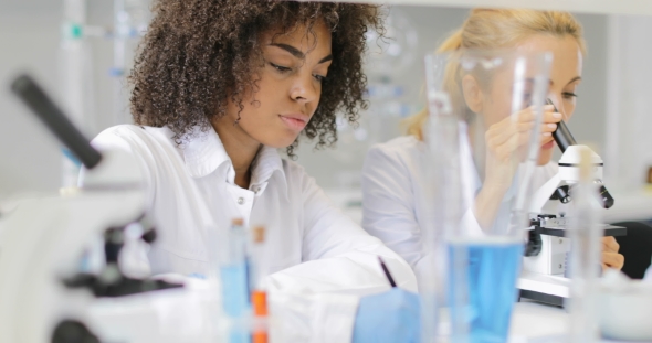 Woman Scientist Working With Microscope While Her African American Colleague Making Notes Of
