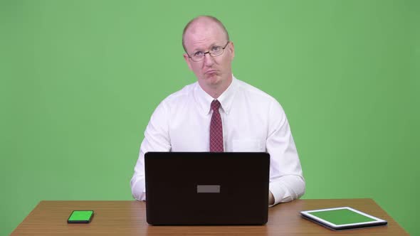 Mature Bald Businessman Using Laptop and Phone at Work Against Wooden Table